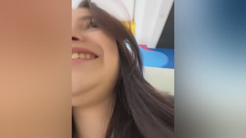 Media: Video of a smiling woman with long dark hair, partially obscured by a large red object on the left and a blue object on the right, in a bright, indoor setting.