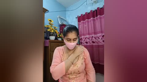 Video of a South Asian woman with medium skin tone, wearing a pink face mask, pink long-sleeve blouse, and beige trousers, sitting on a bed in a modestly decorated room with blue walls, a wooden dresser, and red curtains.