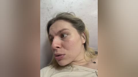 Video of a young Caucasian woman with fair skin, blonde hair, and light makeup, wearing a beige top, standing in a bathroom with a tiled wall. She has a concerned or confused expression.