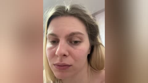 Video of a close-up, fair-skinned woman with shoulder-length blonde hair, wearing minimal makeup, looking slightly down. The background is blurred, with beige and peach tones.