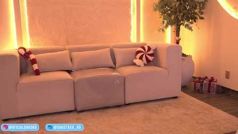 Video of a cozy living room with a beige sectional sofa, festive red and white striped pillow, and a potted tree. Warm ambient lighting and wrapped gifts on the floor.