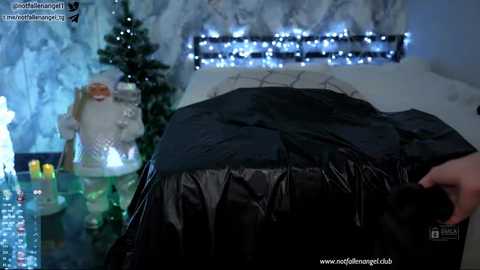 Video of a person with blonde hair, partially covered in black plastic, standing in front of a Christmas tree with a white-bearded Santa, and a snowy, lit-up background.