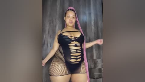 A video of a plus-sized Black woman with light brown skin, wearing a revealing black fishnet bodysuit with cutouts and a pink headscarf. She stands against a wooden wall in a dimly lit room.