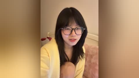 A video of an East Asian woman with straight black hair and glasses, wearing a yellow robe, seated on a bed, holding a baby, with a beige wall background.