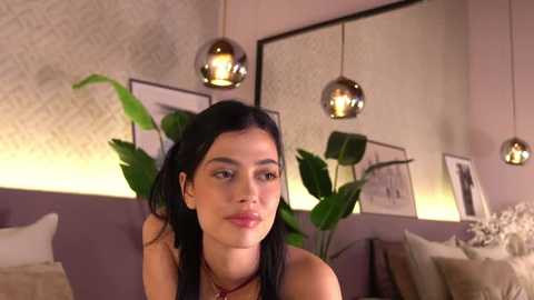 Video of a young woman with dark hair and light skin, wearing a red necklace, in a modern living room with abstract wallpaper, potted plants, and hanging pendant lights.