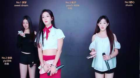 Video of three East Asian women, two with long black hair, in a studio, dressed in white and black, holding microphones, against a dark background.