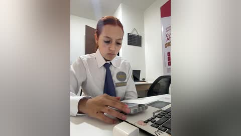 Video of a female office worker with light skin and red hair, wearing a white shirt and blue tie, working on a calculator at a desk.