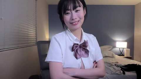 Video of a smiling young Asian woman with dark hair, wearing a white blouse with a large pink bow, standing in a dimly lit bedroom with a bed, lamp, and window.