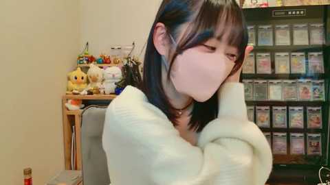 A young woman with straight black hair and light skin, wearing a white sweater, sits with her face covered by a mask, looking down. Behind her is a cluttered room with a toy shelf and a video game console.