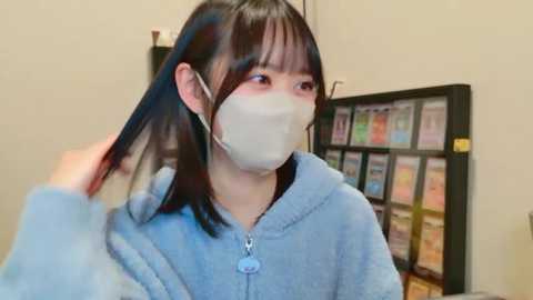 Video of an East Asian woman with shoulder-length black hair, wearing a light blue hoodie and a face mask, adjusting her hair in a public library with colorful bookshelves in the background.