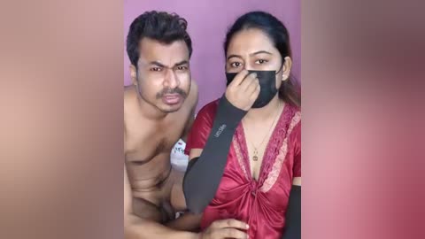 A video shows a muscular, shirtless Indian man with short hair and a beard, and a woman with dark hair and a black face mask, both sitting against a pink background.