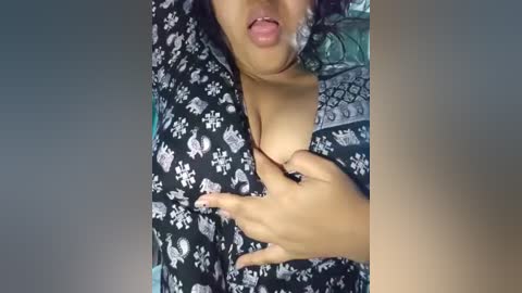 A video of a woman with medium brown skin, wearing a black floral shirt, exposing her large breasts, one hand on her chest. The background is blurred.