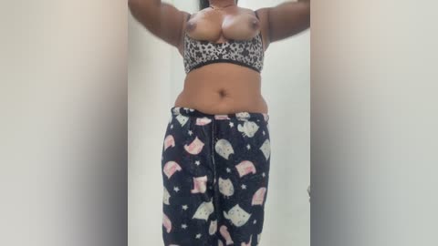 Video of a topless woman with medium brown skin, wearing a leopard print bra and black pajama pants adorned with snowmen, standing against a plain white wall.