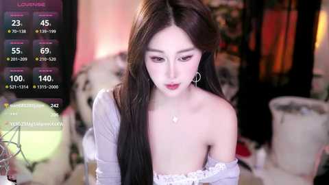 Video of a young East Asian woman with long black hair, wearing a white off-shoulder top, seated in a dimly lit room, with a live stream overlay displaying viewers and chat.