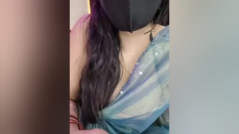 Video of a woman with long, wavy black hair, wearing a black face mask, a blue and white tie-dye shirt, and a black bra strap. The background is blurred, focusing on her upper body.