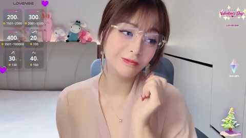 Video of a young Asian woman with light skin, brown hair, and glasses, wearing a beige top, smiling while touching her nose, surrounded by plush toys in a cozy, well-lit room.