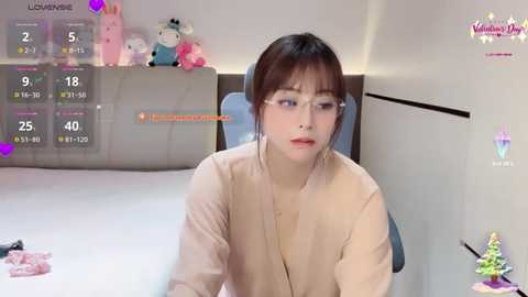 Media: A photo of a young East Asian woman with fair skin and dark hair, wearing glasses and a beige top, sitting on a bed with plush toys and a calendar.