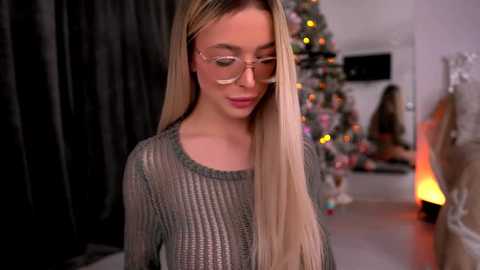 Video of a young, slender blonde woman with fair skin, wearing glasses and a sheer, grey knit sweater, standing in a cozy, dimly-lit living room with a decorated Christmas tree and fireplace in the background.