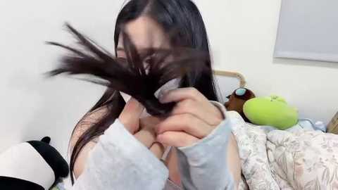 Media: A video of a woman with long black hair, wearing a light blue sweater, playfully tugging her hair. The background shows a white wall, a bed with a green stuffed animal, and a window with a white blind.