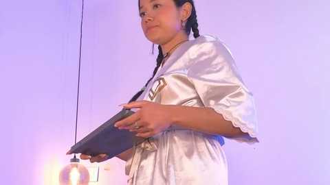 Video of a young woman with medium brown skin and braided hair, wearing a white satin robe, holding a black tablet, against a soft purple background.