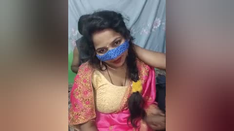 Video of an Indian woman in traditional pink and gold saree, wearing a blue face mask, with a man's arm around her shoulder. Background shows a gray curtain with floral patterns.