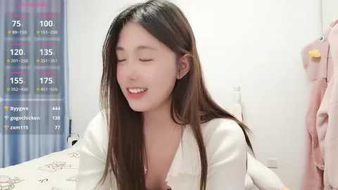 A video of a young East Asian woman with long black hair, smiling, wearing a white blouse, in a bedroom with a plaid curtain and pink towel.