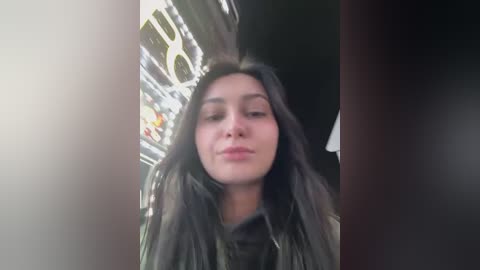 Video of a young woman with long, straight black hair, wearing a black leather jacket, standing in a dimly lit, narrow hallway. Her face is slightly tilted, with a neutral expression.