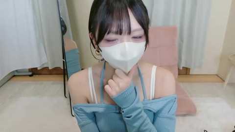 Video of an Asian woman with dark hair, wearing a white face mask, a blue off-shoulder sweater, and a white bra. She kneels on a light carpet in a room with beige walls and a pink chair.