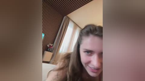 Video of a young woman with long brown hair, fair skin, and green eyes, smiling slightly, indoors with striped wallpaper, curtains, and a beige couch in the background.