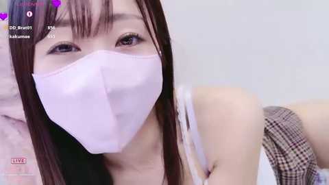 Video of an East Asian woman with long, straight black hair and fair skin, wearing a white mask and plaid skirt, in a bedroom with pink bedding.