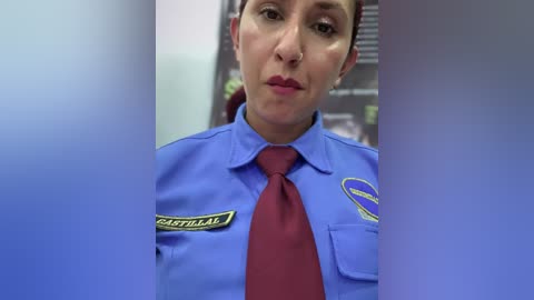 Video of a Latina woman with short hair, wearing a blue police uniform with a red tie, badge, and nameplate, standing in a blurred background.
