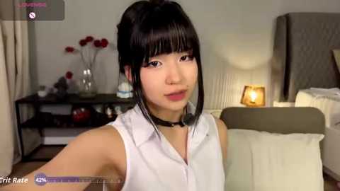 Media: A video of an Asian woman with straight black hair in a ponytail, wearing a white blouse and black choker, sitting in a modern bedroom with beige walls, a bed, and a lit lamp.