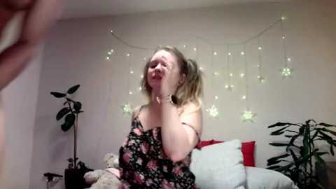 Video of a smiling woman in a black floral dress, sitting on a bed with plush toys and a potted plant, against a background of fairy lights.
