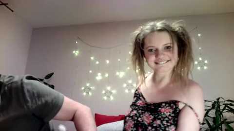 Video of a young, fair-skinned woman with light brown pigtails, smiling, wearing a black floral dress, in a cozy room with string lights and a potted plant in the background.