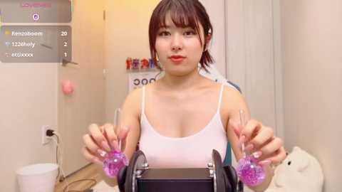 Video of an East Asian woman with short dark hair and red lipstick, wearing a white tank top, sitting in a clinic room with a black machine and purple plugs in her ears.