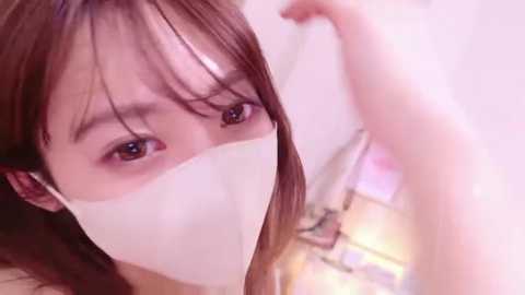 Video of a young Asian woman with straight, light brown hair and large, brown eyes, wearing a white face mask. She is indoors, possibly in a bedroom, with soft, pastel-colored walls and a faintly visible bed in the background.