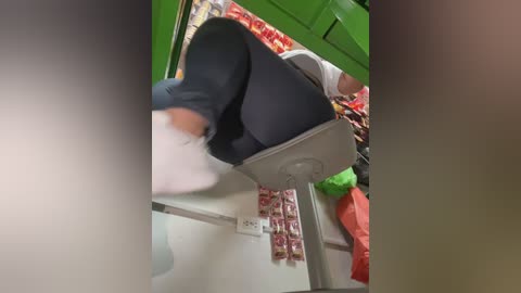 Video of a woman in a white shirt and black leggings, seated on a gray chair, inside a brightly lit store with green walls and red and yellow merchandise on shelves.