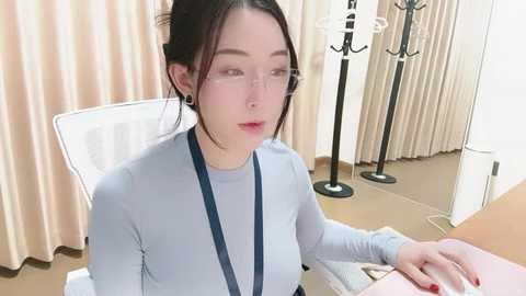 A video of a fair-skinned, young Asian woman with dark hair tied back, wearing glasses and a light blue long-sleeved top, seated at a desk, typing on a pink laptop. The background features beige curtains and medical equipment, suggesting a hospital or clinic setting.