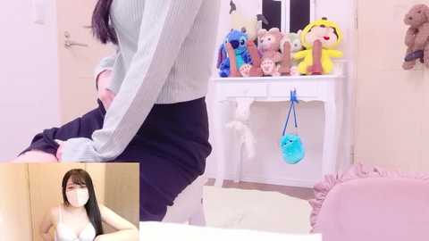A video of a young woman with long black hair and glasses, topless, in a room with stuffed animals, plush toys, and a white vanity table.
