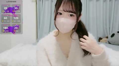 Video of a young East Asian woman with light skin, brown hair, and bangs, wearing a white fluffy robe and mask, standing in a bedroom with a gray curtain and a stuffed animal.