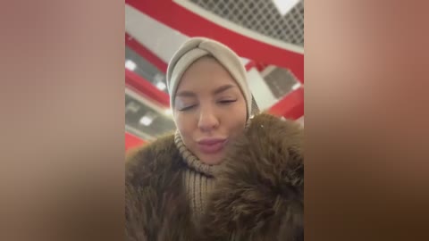 A video of a young woman with fair skin, wearing a beige beanie, brown fur coat, and a beige turtleneck sweater, puckering her lips, against a blurred, red-and-white background, possibly a concert venue.
