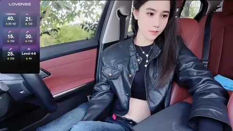 Media: Video of a young Asian woman with long dark hair, wearing a black leather jacket and jeans, sitting in a car with a digital display showing location and time.