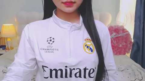 Video of a woman with long black hair, wearing a white Emirates FC soccer jersey, standing in a softly lit bedroom with a bed, lamp, and red heart-shaped pillow.