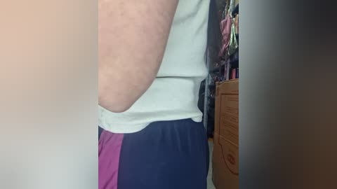 Video of a person wearing a white tank top and dark blue shorts, with a visible tattoo on their left arm, standing in a cluttered room with a wooden dresser and clothes hanging in the background.