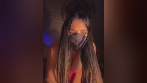 Video of a woman with long, straight, dark hair wearing a black mask, pink top, and a large, dark hat, blurred background with dim lighting.