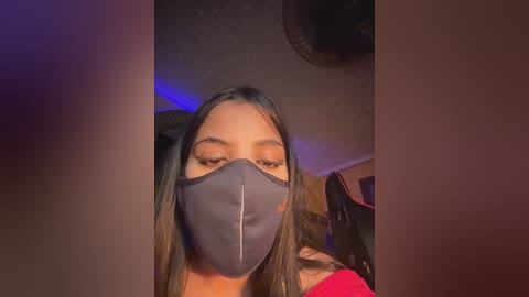 Video of a young woman with long, straight black hair, wearing a dark blue surgical mask and a red top, indoors with dim lighting and a ceiling fan visible.