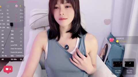 Media: A video of a young Asian woman with straight black hair, wearing a light blue tank top, sitting on a white couch in a cozy room.