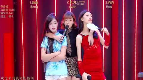 Video of three women, one with a microphone, standing on stage with red curtains in the background.