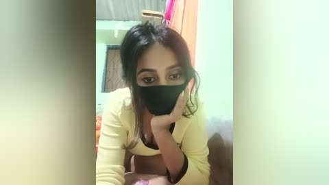 Media: Video of a young woman with long dark hair, wearing a yellow top and black face mask, taking a selfie in a dimly lit room with a wooden door and hanging clothes in the background.