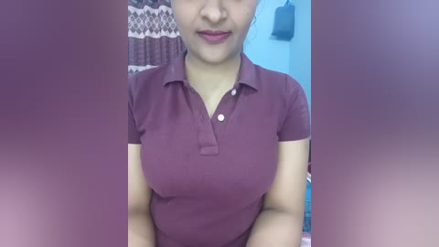 Media: A video of a woman with medium skin tone, wearing a purple polo shirt, standing in a bathroom with a patterned shower curtain and a blue towel rack.